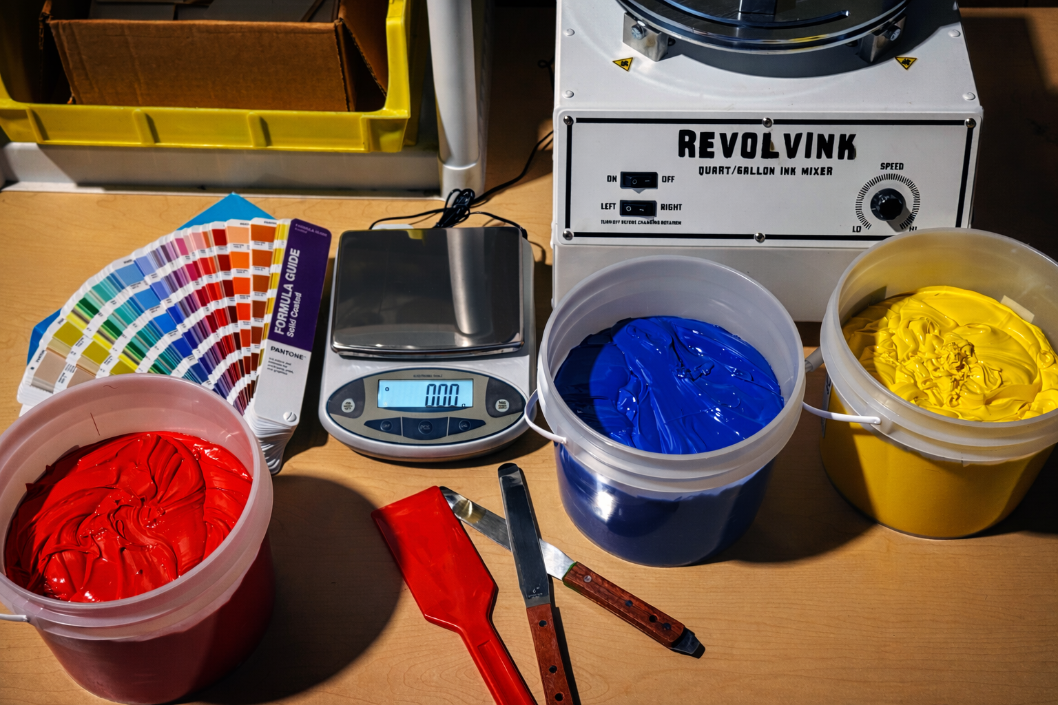 Color swatch, digital scale, and buckets of red, blue, and yellow ink on a table with Revolvink equipment.